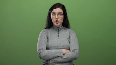 Esmer, gözlüklü bir kadın yeşil arka plan kollarına yaslanmış, neşeli bir şekilde somurtuyor gri kazak giyiyor.