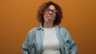 Orta yaşlı, kıvırcık saçlı ve gözlüklü bir İspanyol kadın canlı turuncu bir arka plana karşı aptal bir yüz ifadesi takınıyor. Sıradan bir kot gömlekle neşeli ve komik titreşimler yayıyor..