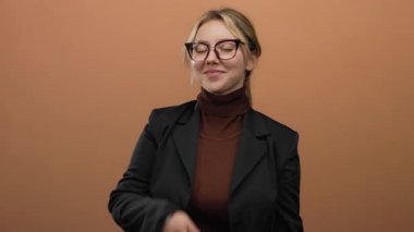 Kadın parmaklarıyla zafer işareti yaparken gülümsüyor, kahverengi bir duvara yaslanıyor, gözlük ve ceket takıyor, kendinden emin ve neşeli bir ifade sergiliyor..