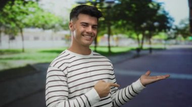 Parkta gülümseyen, çizgili tişörtlü, açık havada yemyeşil bir ortamda, davetkar ve neşeli bir atmosferi canlandıran genç bir adam..