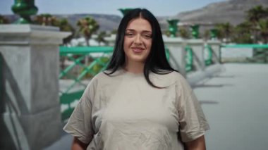 Uzun siyah saçlı bir kadın, yeşil korkuluklar ve güneşli arka planda palmiye ağaçları olan bir caddede dururken kameraya öpücük yolluyor..