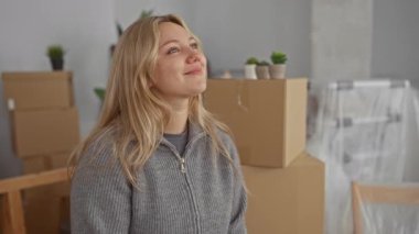 Yeni bir evde gülümseyen bir kadın. Oturma odasındaki kutuları taşıyarak yeni bir başlangıç umudu ve heyecanı simgeliyor..