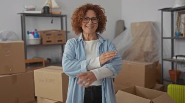 Oturma odasında gülümseyen kahverengi saçlı bir kadın taşınan kutularla çevrili yeni bir ev ortamında raflarla ve arka planda paketlenmemiş eşyalarla yeni bir başlangıç yaptığını gösteriyor..