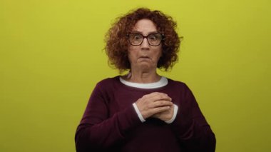 İspanyol orta yaşlı, kıvırcık saçlı, gözlüklü bir kadın canlı sarı izole bir arkaplan karşısında korkmuş görünüyor. İfadesinde duygu ve yoğunluk gösteriyor..