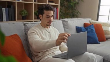 Genç adam rahat oturma odasında dizüstü bilgisayarda çalışıyor, renkli yastıklar ve modern dekorlarla çevrili, rahat ve odaklanmış bir ofis ortamını öneriyor..