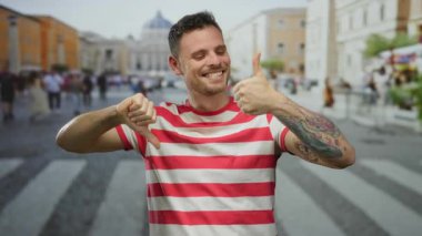 Çizgili gömlekli genç adam şehir sokaklarında karışık başparmak hareketleri ve şehir ortamında dövmeli kol gösteriyor..