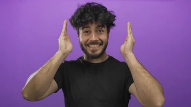 Sakallı genç İspanyol bir adam, parlak mor bir arka plana karşı elleriyle yüzünü örtüp açarken ciddi ve gülümseyen yüz ifadeleri arasında gidip geliyor..