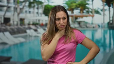 Lüks bir otelde, havuz başında boğazını tutmaktan rahatsız olan bir kadın. Pembe bir gömlek giyiyor, açık havada sağlık ve sağlığın temasını resmediyor..