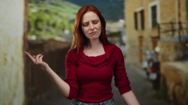 Kırmızı tişörtlü, kırmızı saçlı bir kadın şehir sokaklarında dalgın ve çekici bir şekilde dikiliyor. Bulanık binalarla sakin bir şehir ortamı sergiliyor..