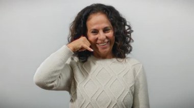 Kıvırcık saçlı ve neşeli suratlı bir kadın beyaz arka planda telefon jesti yapıyor..