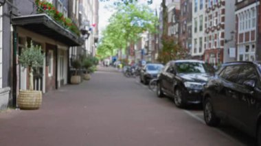 Amsterdam 'daki bir Avrupa sokak manzarasının bulanık görüntüsü, park halindeki arabaların tarihi binalar ve yeşillik çerçeveli tarihi bir kaldırım yolu ve mavi gökyüzü altında.