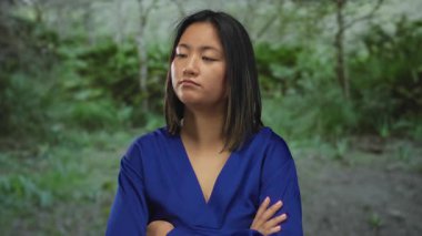 Parkta kolları çapraz duran mavi bluz giyen bir kadın, etrafında bulanık yeşil yeşillik ile doğal bir ortamda kendine güvenini ifade ediyor, düşünceli ve huzurlu görünüyor..