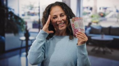 Orta yaşlı bir kadın elinde yeni Zelanda banknotları tutarken gülümsüyor. Bir otel resepsiyonunda, zenginlik ve finansal başarı sergiliyor..