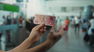 Arka planda bulanık uçuş kalabalığı, seyahat ve finans konularını vurgulayan Türk lira banknotunu havaalanında tutan adam.