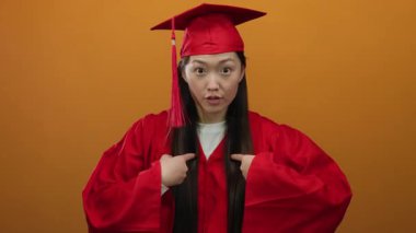 Kırmızı mezuniyet elbisesi ve şapkalı Asyalı kadın turuncu arka plan jestlerine karşı poz veriyor. Her iki eli de başarıyı içten kutluyor ve başarının gururunu dile getiriyor..