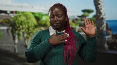 Pembe örgülü bir kadın, yeşil bir kazakla açık havada, yemyeşil bir parkın arka planına karşı yemin jesti yapıyor. Bağlılığını ve samimiyetini vurgulayan resmi bir anı yakalıyor..