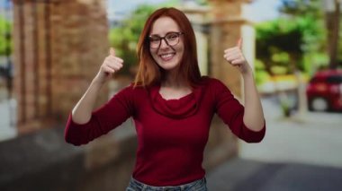 Şehrin bir caddesinde başparmağıyla gülümseyen bir kadın kızıl saçlı ve gözlüklü, mutluluk, neşe ve pozitiflik taşıyor..