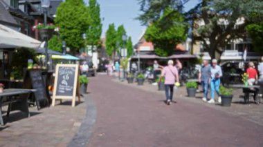 Güneşli bir günde arka planda güneşli şehir yaşamını yansıtan yemyeşil alanlar ve açık kafelerle Avrupa 'nın büyüleyici caddesinde rahatça yürüyen insanlar.