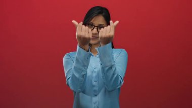 Gözlüklü bir kadın kalp hareketi yapıyor parmaklarıyla izole edilmiş kırmızı bir arkaplanı işaret eden mavi bir gömlek giyiyor. Bağlantı ve sıcaklığı gösteriyor..