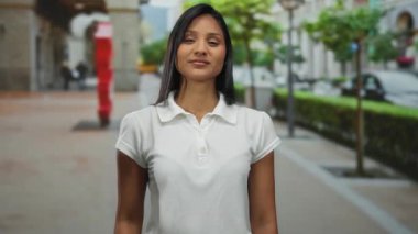 Güneşli bir şehir sokağında, arka planı bulanık bir şekilde kameraya doğru uzanan bir kadın açık bir şehir ortamında kendinden emin ve misafirperver bir jest sergiliyor..