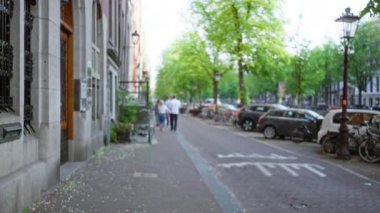 Amsterdam 'da bulanık bir şehir manzarası. Odaklanamayan yayalar, park halindeki arabalar ve tarihi mimariyle çevrili ağaçlık bir cadde boyunca yürüyor..