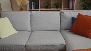 Modern oturma odasındaki kanepede oturan genç adam rahat ev ortamında rahat giysiler giymiş arka planda kitaplar ve bitkiler sergileniyor rahat bir ev hayatı sergiliyor.