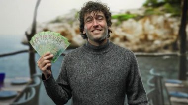 İspanyol adam restoran terasında neşe içinde Rus rubleleri sergiliyor. Şehir sokaklarında para ve mutluluğun özünü yakalıyor..