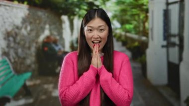 Kentsel sokak ortamında gülümseyen bir kadın pembe giyinmiş, yeşillikle mutluluğu ifade eden ve parlak bir günde bulanık arka planda oturan insanlar..