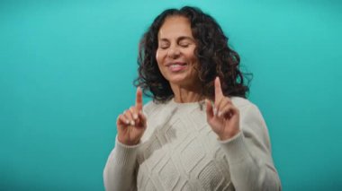 Kıvırcık saçlı olgun İspanyol kadın canlı mavi bir arka planda neşeyle dans ediyor, mutluluğu ve tasasız ruhu ifade ediyor..