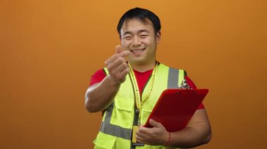 Yelekli genç adam turuncu arka plana kalp hareketi yapıyor..