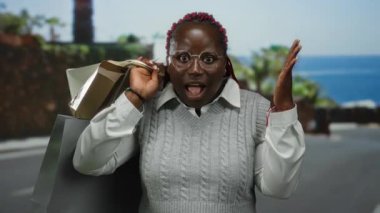 Kumsal girişine şaşıran bir kadın, güneşli bir günde elinde okyanus ve palmiye ağaçlarıyla birlikte açık havada alışveriş torbaları tutuyor..