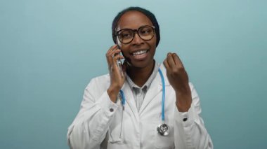 Akıllı telefondan konuşan bir kadın, boynunda steteskopla, izole edilmiş mavi bir arkaplan ile konuşuyor. Duygularını jestlerle ifade ediyor ve gözlük takıyor..
