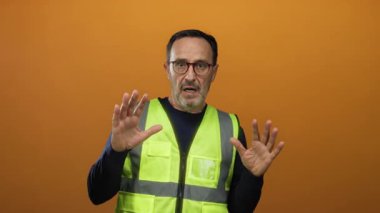 Güvenlik yeleği giyen İspanyol bir adam parlak turuncu duvar arkasına şaşırmış görünüyor, aciliyet ve tetikte olma duygusunu somutlaştırıyor..