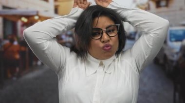 Gözlüklü bir kadın şehir terasında, canlı bir şehir ortamında neşeli bir ifade sergileyen tavşan kulaklı bir jest yapıyor..