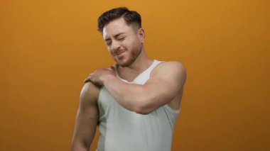 Beyaz kolsuz tişörtlü genç adam izole edilmiş turuncu arka plana karşı omuz ağrısı çekiyor. Rahatsızlık ve sağlık teması taşıyor..