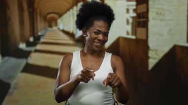 Tarihsel üniversite koridorunda gülümseyen ve işaret eden bir kadın canlı akademik ortam sergiliyor..