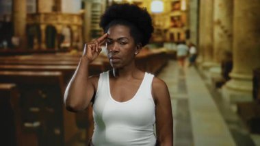 Kilisede duran bir kadın, arka planda bulanıklığa karşı düşünceli bir ifadeyle kafasına dokunuyor, beyaz atlet giyip iç mekan ayarları yapıyor..