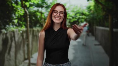 Güneşli bir şehir caddesinde gülümseyen kızıl saçlı kadın yeşil ağaçlarla kameraya doğru uzanarak canlı ve canlı bir zemin oluşturuyor..
