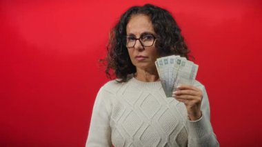 Orta yaşlı, gözlüklü, elinde Mısır banknotları olan ve el kol hareketi yapan bir kadın cesur bir kırmızı arka planda durup ekonomik uyarı ve güçlü bir finansal görüş mesajı iletiyor..