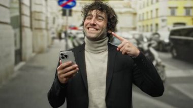 Kıvırcık saçlı, gülümseyen ve telefonuna bakan genç bir adam işlek bir caddede, şehir tarzını somutlaştıran ve açık hava ortamında gençlik enerjisi veren..