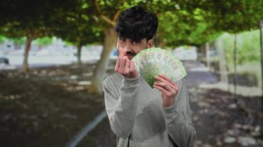 Parkta elinde Rus rublesi banknotlarıyla gülümseyen bir adam, çevresi yeşilliklerle çevrili, açık hava zenginliği ve finans kavramını tasvir ediyor..