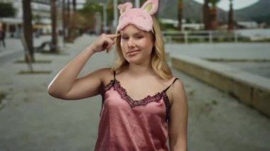 Kumsalda gece elbisesi giymiş genç sarışın bir kadın gündüz eğlencesine pembe uyku maskesi takmış komik suratlar yapıyor..