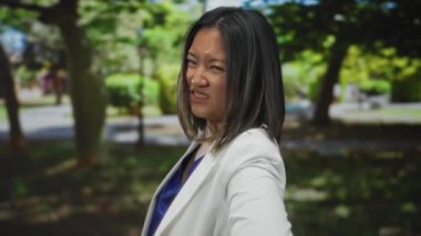 Parkta beyaz ceketli genç Çinli kadın, sırt ağrısını ifade ediyor, parlak bir gökyüzünün altında yemyeşil ağaçlarla çevrili, huzursuzluk ve doğanın huzursuzluğunu gösteriyor..