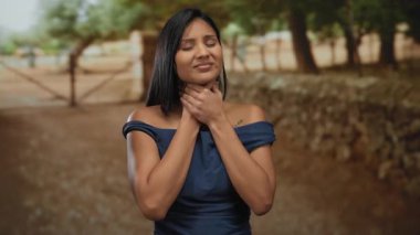 Mavi elbiseli bir kadın parkta boğazını tutuyor ve yeşil ve gün ışığında rahatsız görünüyor..