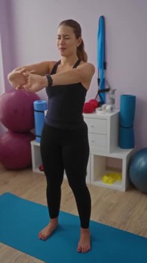 Kadın kapalı alanda egzersiz yapıyor, spor salonundaki mavi yoga minderinin üzerinde spor aletleri ile geriniyor, dingin bir spor ortamında odak ve dengeyi gösteriyor..