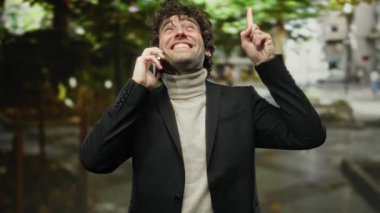 Genç adam canlı bir şehir caddesinde telefonla konuşurken mutlu bir şekilde yukarıyı işaret ediyor..