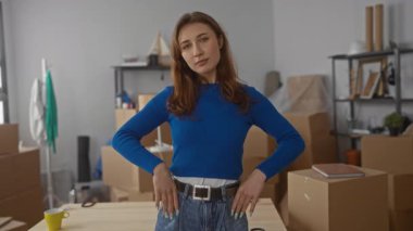 Mavi kazaklı kadın, yakın zamanda satın aldığı apartmanın içinde yeni bir başlangıcı temsil eden taşınan kutularla çevrili modern oturma odasında kollarını kavuşturuyor..