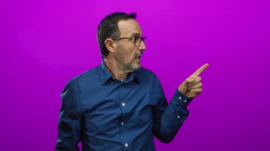 Gözlüklü olgun bir adam mavi gömlekli canlı pembe bir arka plana hevesle bakıyor..