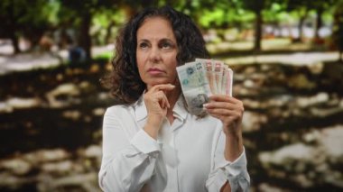 İngiliz sterlini tutan bir kadın, bir park ortamında düşünceli bir şekilde, güneş ışığı altında düşünce ve karar verme duyusunu iletiyor..
