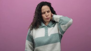 Üzüntülü bir ifadeyle pembe arka planda duran bir kadın, sanki rahatsız olmuş gibi boynunu tutuyor, çizgili bir kazak giyiyor, stresi veya gerginliği vurguluyor.